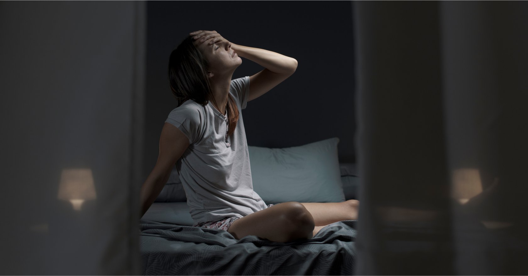 woman sitting on bed in dark bedroom holding her head in exhasperation